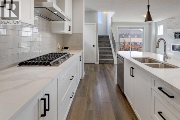 View of a white kitchen with a gas stovetop, island, and staircase leading to the upper level. - 112 Emberside Place, Cochrane