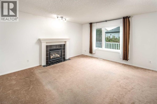 Living room with a beige carpet, white walls, and a fireplace with dark tiles. - 112 Everridge Drive Sw, Calgary