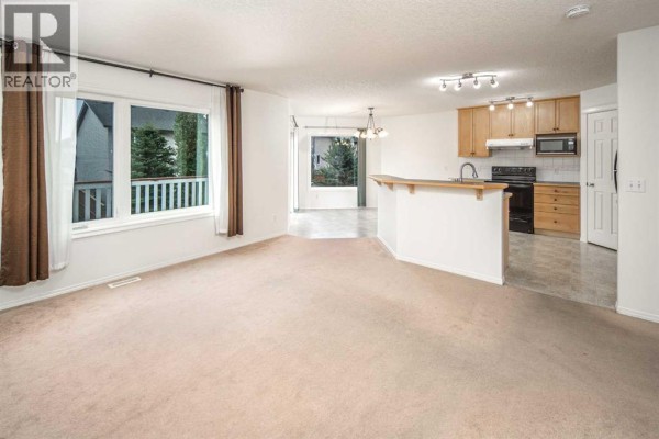 Living room and kitchen area with beige carpet, a breakfast bar, and light wood cabinets. - 112 Everridge Drive Sw, Calgary