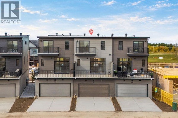 Rear exterior view of townhouses with balconies, garage doors, and grey siding. - 112 Greenwich Drive Nw, Calgary