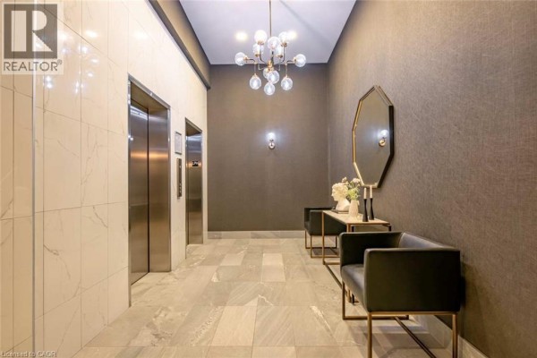 Building hallway with elevators, modern lighting fixture, decorative mirror, and seating benches. - 112 King Street E Unit# Lph01, Hamilton