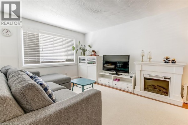 Living room with a grey sectional sofa, white fireplace, television, and blinds on the window. - 112 Richardson Drive Unit# 24, Port Dover
