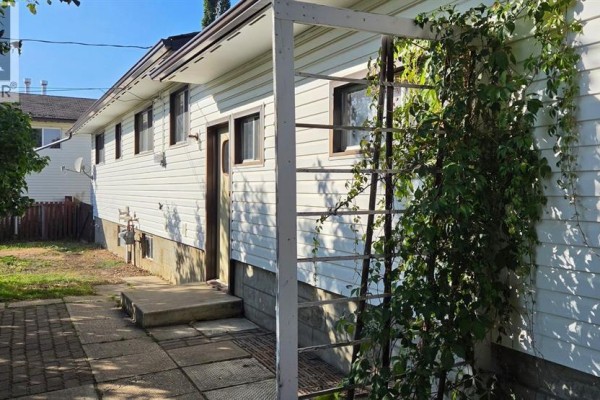 Side exterior of a white vinyl-sided house with a brick pathway and a trellis with vines. - 11221 102 Avenue, Fairview