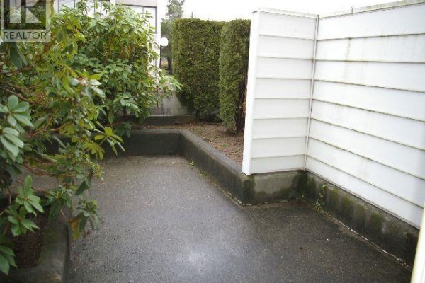 A paved patio is surrounded by concrete borders, green bushes, and a white vinyl fence. - 113 707 8 Street, New Westminster