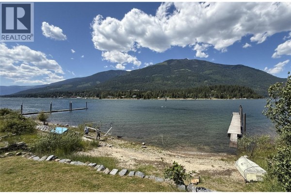 View of Kootenay Lake and mountains from a grassy shoreline with a dock. - 113 Kootenay Lake Road, Procter