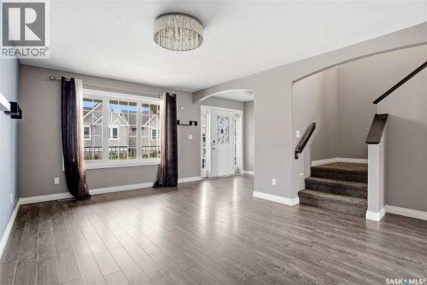 Living room with wood-look flooring, large window, and staircase leading upstairs. - 1137 Jurasin Street N, Regina