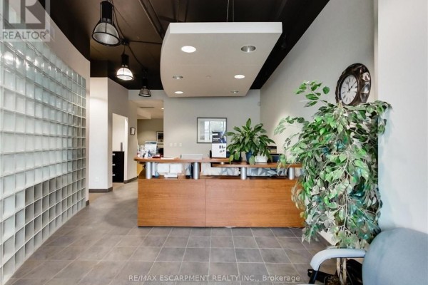 Reception desk with wooden counter, black ceiling lights, and a large plant next to a wall clock. - 114 - 1100 South Service Road, Hamilton
