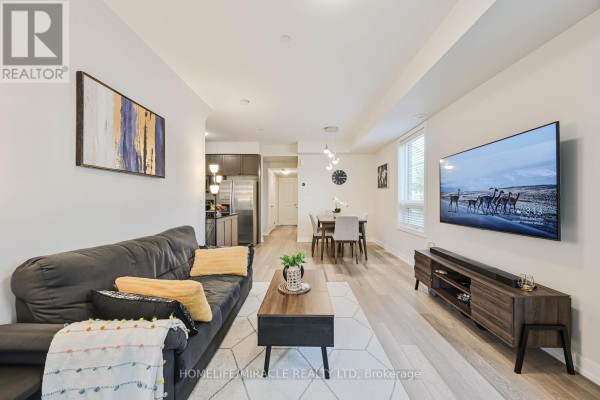 Living and dining room with dark sofa, dining table, and kitchen in background. - 114 - 256 Royal York Road, Toronto