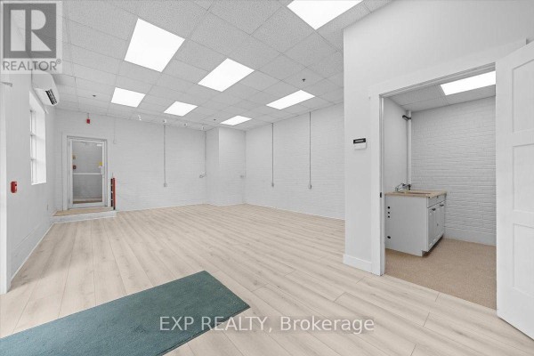 Empty commercial interior with white brick walls, light wood laminate flooring, drop ceiling with fluorescent lights, and a small washroom area. - 114 - 40 Sunset Boulevard, Perth