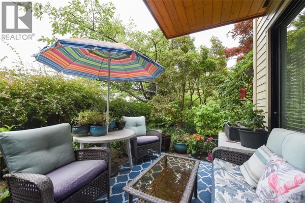 Patio with wicker chairs, a table, an umbrella, and dense green foliage. - 114 2300 Mansfield Dr, Courtenay