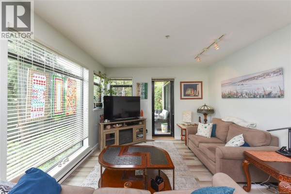 Living room with a large window, sectional sofa, coffee table, television, and a sliding door to a patio. - 114 2300 Mansfield Dr, Courtenay