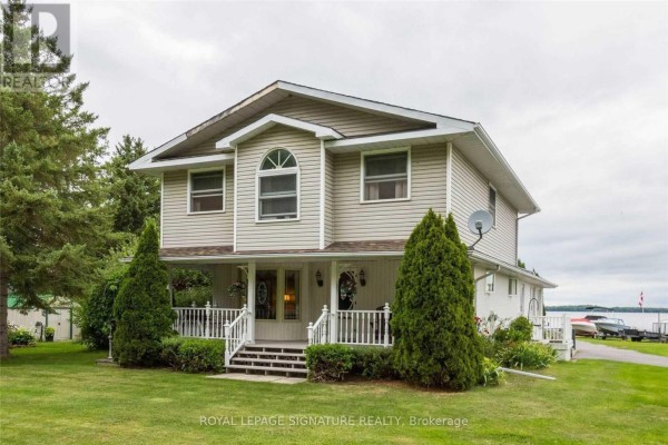 Two-story beige house with white railings on the porch, surrounded by green lawn and trees. - 11438c Loyalist Parkway, Prince Edward County