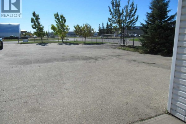 Paved lot with a chain-link fence bordering trees and industrial buildings in the distance. - 11450 89 Avenue, Grande Prairie