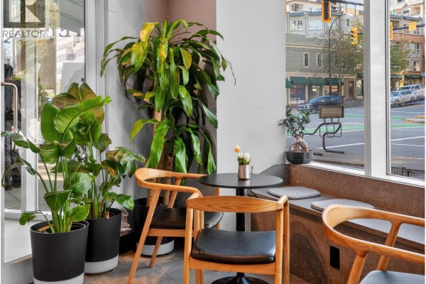 Corner seating in a cafe with large potted plants, two small tables, and windows showing a street and buildings. - 11450 Confidential, North Vancouver
