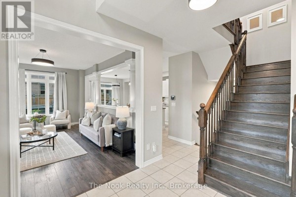 View from the foyer into a bright living room with light gray walls, white furniture, and a dark wood staircase. - 115 Noden Crescent, Clarington