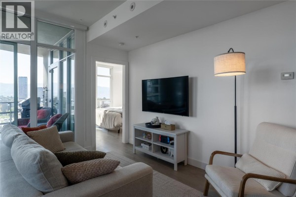 Living room with a sectional sofa, armchair, television, and a view of a bedroom through an open doorway. - 1151 Sunset Drive Unit# 1305, Kelowna