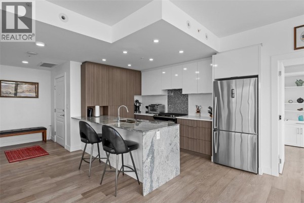 Modern kitchen with a large island, granite countertops, white and wood cabinets, and stainless steel appliances. - 1151 Sunset Drive Unit# 1504, Kelowna