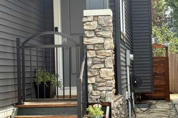 Close-up of front porch stairs with stone pillar, black metal railing, and potted plants. - 1155 Kings Heights Way Se, Airdrie