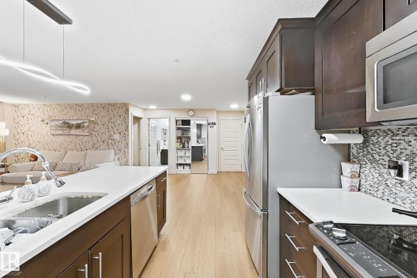 Kitchen island with seating, dark cabinets, and view into living room with patterned wallpaper. - #116 4008 Savaryn Dr Sw Sw, Edmonton
