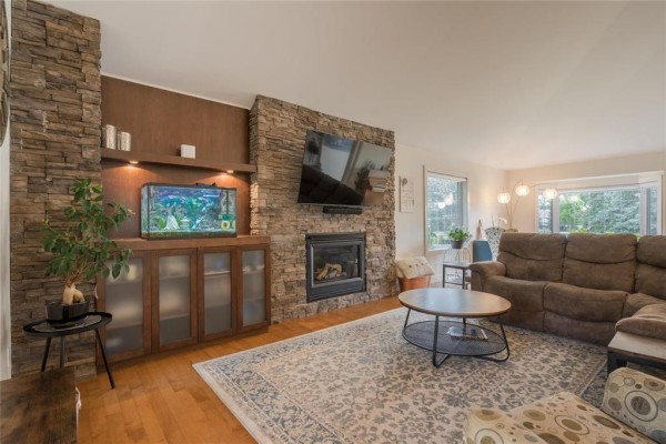 Living room with a stone fireplace, mounted TV, aquarium, wood-paneled wall, and hardwood floors. - 116 Harvest Drive, Steinbach