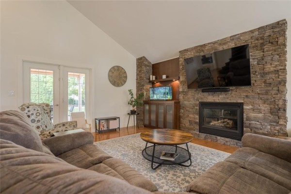 Living room with a stone fireplace, hardwood floors, a large sectional couch, and double glass doors. - 116 Harvest Drive, Steinbach