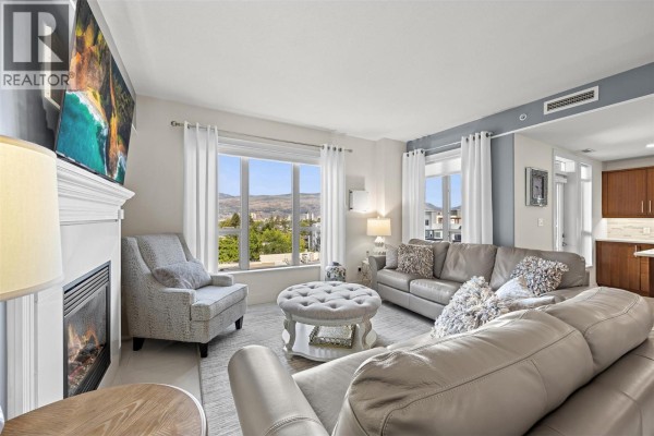 Living room with gray couches, fireplace, armchair, ottoman, and large window. - 1160 Bernard Avenue Unit# 601, Kelowna