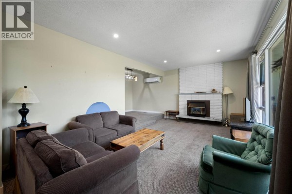 Living room interior with a white brick fireplace, beige carpet, and large window. - 1160 Kemano Street, Kamloops
