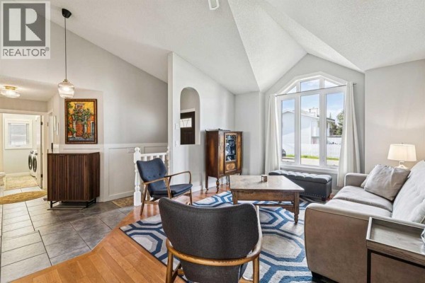 Living room with wood flooring, vaulted ceiling, arched window, sofa, armchairs, and a view into the foyer. - 117 Hawkbury Close Nw, Calgary