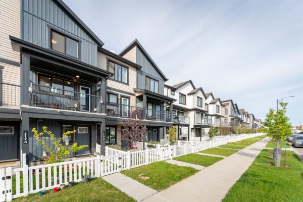 Street view of townhouses with beige and dark gray siding, balconies, and white picket fences along a sidewalk. - 1172 Aster Boulevard Nw Nw, Edmonton