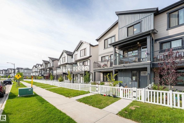 View down the street of multiple townhouses with varying colors and balconies, featuring white picket fences and green lawns. - 1172 Aster Boulevard Nw Nw, Edmonton