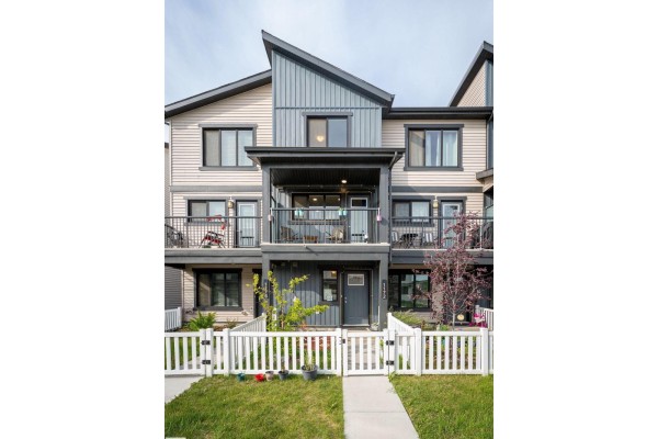 Close-up exterior shot of a townhouse unit with dark gray siding, a covered balcony, and a white picket fence. - 1172 Aster Boulevard Nw Nw, Edmonton
