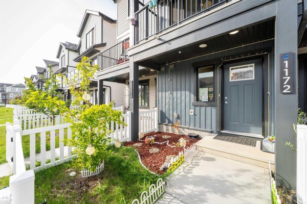 Close-up exterior view of a townhouse entrance with dark gray siding, a gray door, and a small landscaped garden. - 1172 Aster Boulevard Nw Nw, Edmonton