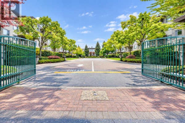 Open green gates reveal a paved driveway lined with trees and hedges. - 118 8180 Jones Road, Richmond