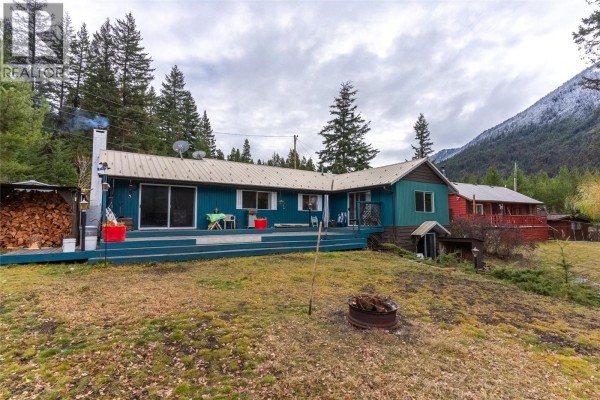 Blue ranch-style house with a large wooden deck overlooking a grassy yard and forest. - 11805 Fountain Valley Road, Lillooet