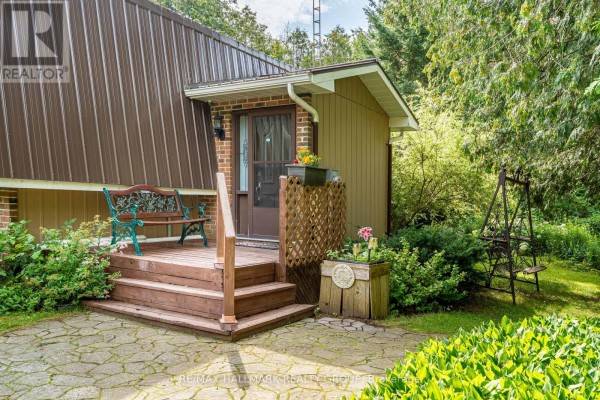 Close-up of a wooden porch with a bench and door, surrounded by lush landscaping. - 1184 Derry Side Road, Beckwith