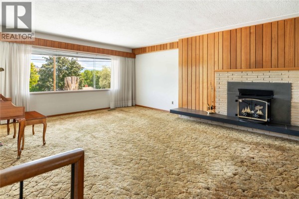 Living room with beige carpet, a brick fireplace, wood paneled walls, and a large window view. - 1188&1190 Hartwick Street, Kelowna