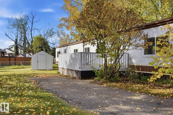 Side view of a white manufactured home with a deck and a gravel driveway surrounded by grass and trees. - 119 Parkland Acres, Lacombe