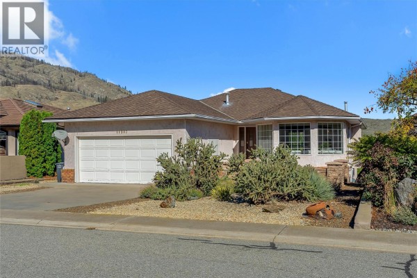 Front of a residential house with a double garage door and a bay window, surrounded by desert-style landscaping. - 11907 Quail Ridge Place, Osoyoos