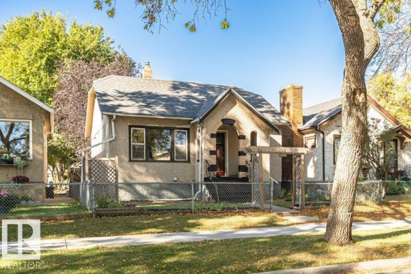 Side view of the beige stucco house with a chain-link fence and mature trees. - 11910 94 St Nw, Edmonton