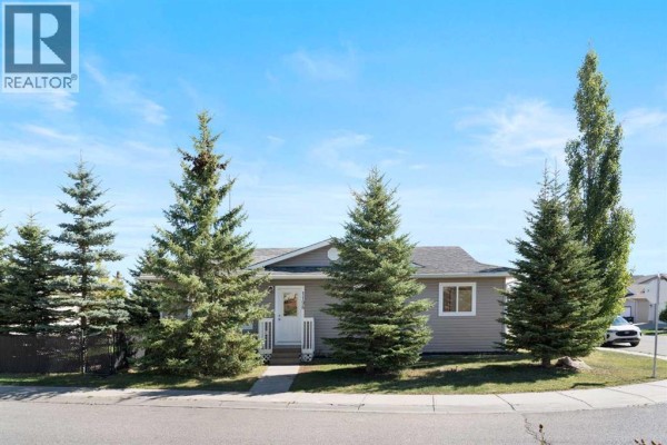 Wide view of the front exterior of a single-story beige house with pine trees. - 1198 Kildeer Close, Didsbury