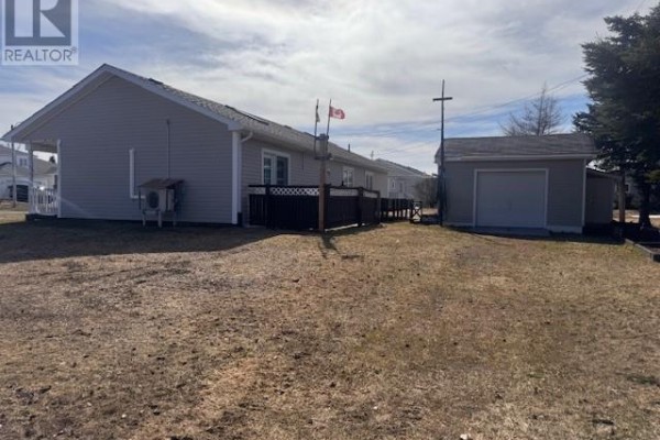 Rear view of the light grey bungalow showing a wooden deck, detached garage, and a large grassy backyard. - 12 Cedar Drive, Lewisporte