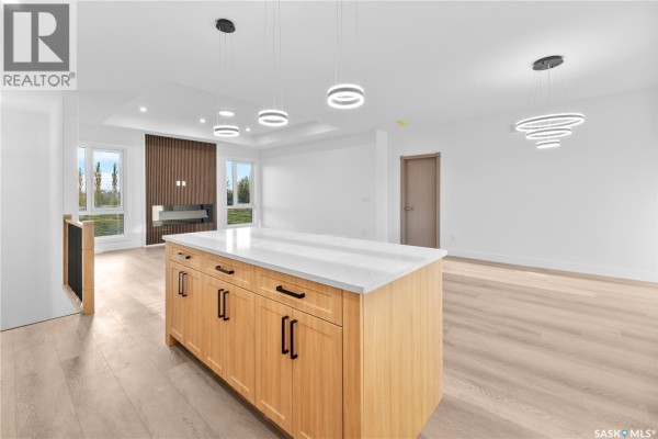Living room with a modern fireplace, wood accent wall, large windows, and a central kitchen island. - 12 Elkwood Cove, Dundurn Rm No  314