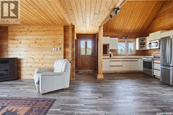 Living area with a white armchair, wood-paneled walls and ceiling, and an open-plan kitchen with white cabinets. - 12 Finch Crescent, Thomson Lake