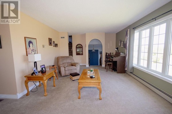 Living room with tan walls, large bay window, piano, and beige carpet. - 12 Hancott's Lane, Frederickton