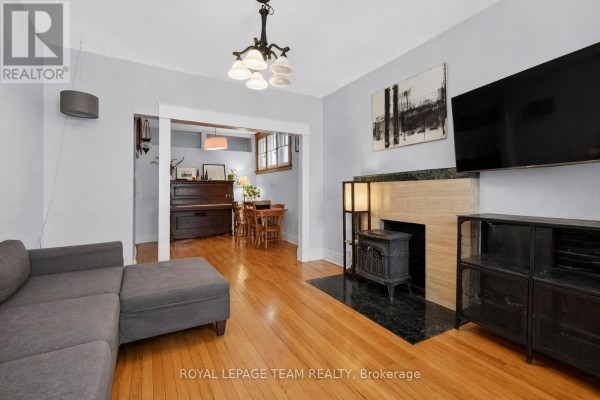 Living room with gray sofa, fireplace, wood floors, and view into a dining area with a piano. - 12 Lewis Street, Ottawa