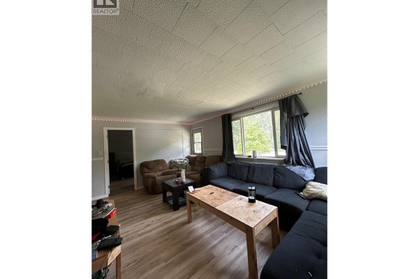 Spacious living room with a large sectional sofa, coffee table, and wide window overlooking greenery. - 1201 Victoria Road W, Revelstoke