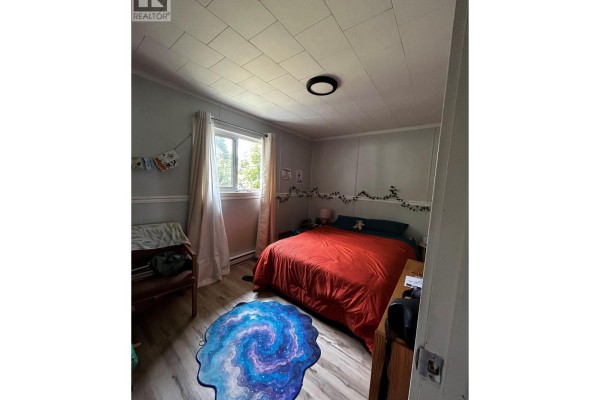 Cozy bedroom featuring a queen-size bed with red bedding and a window with sheer curtains. - 1201 Victoria Road W, Revelstoke