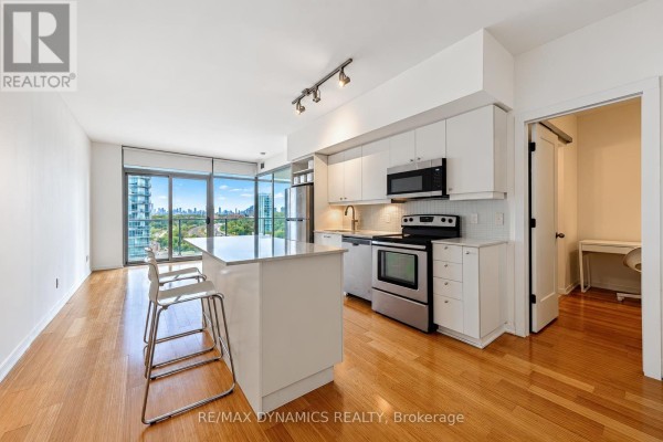 Open-concept living space with a kitchen island, chairs, and a city view through large windows. - 1202 - 103 The Queens Way, Toronto
