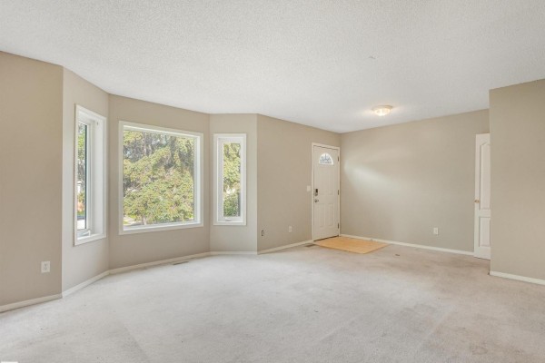 Living room with bay windows facing trees, beige carpet, and a front door. - 12027 105 St Nw, Edmonton