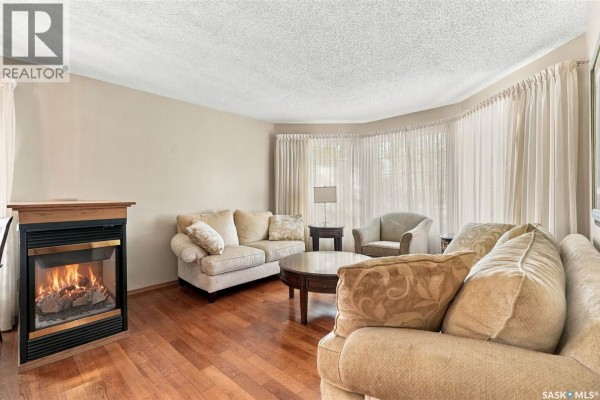 Living room with a gas fireplace, beige sofas, a round coffee table, and large bay windows. - 1204 Lapchuk Crescent N, Regina